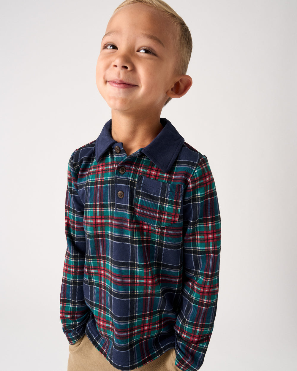 A young boy wearing a long-sleeve plaid button-up shirt in navy blue with red, green, and white stripes, paired with khaki pants, photographed against a light gray studio background.