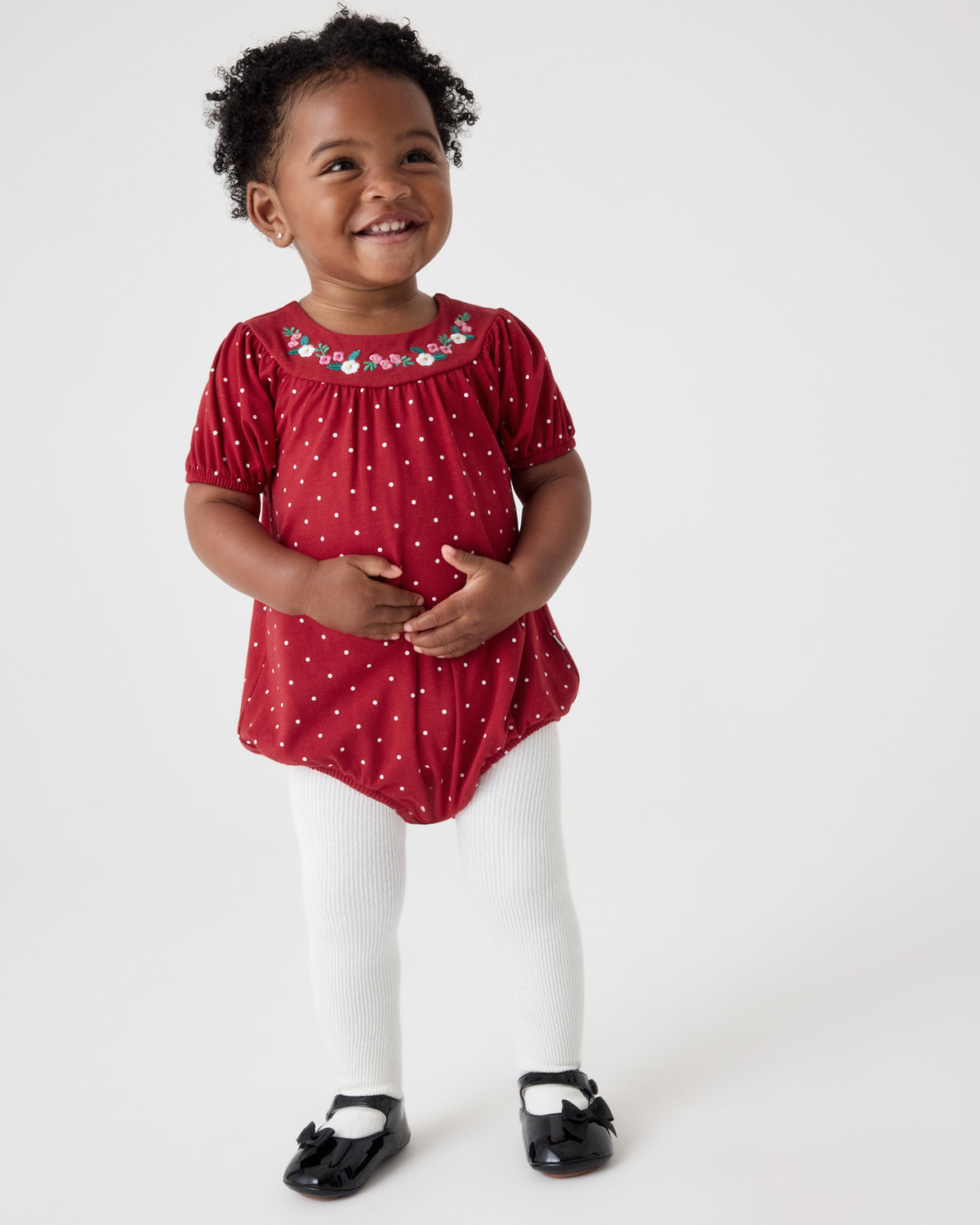 A smiling toddler girl wearing a red short-sleeve top with white polka dots and a decorative embroidered neckline, paired with white tights and black Mary Jane shoes, standing against a plain white studio background.