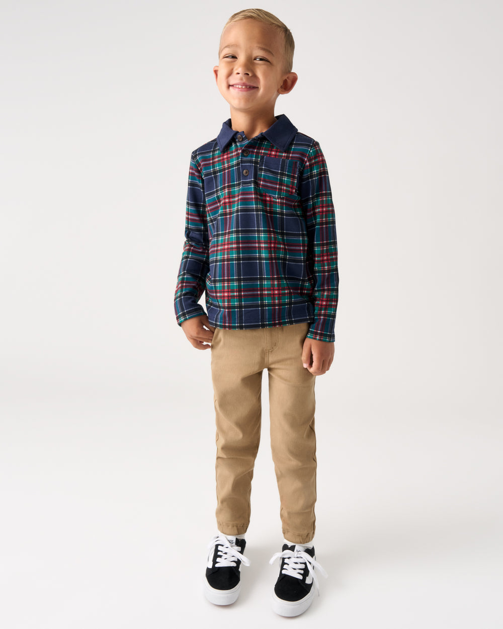 A young boy modeling a long-sleeve plaid button-up shirt in navy blue with red and green stripes, paired with tan pants and black sneakers, standing against a plain white studio background.