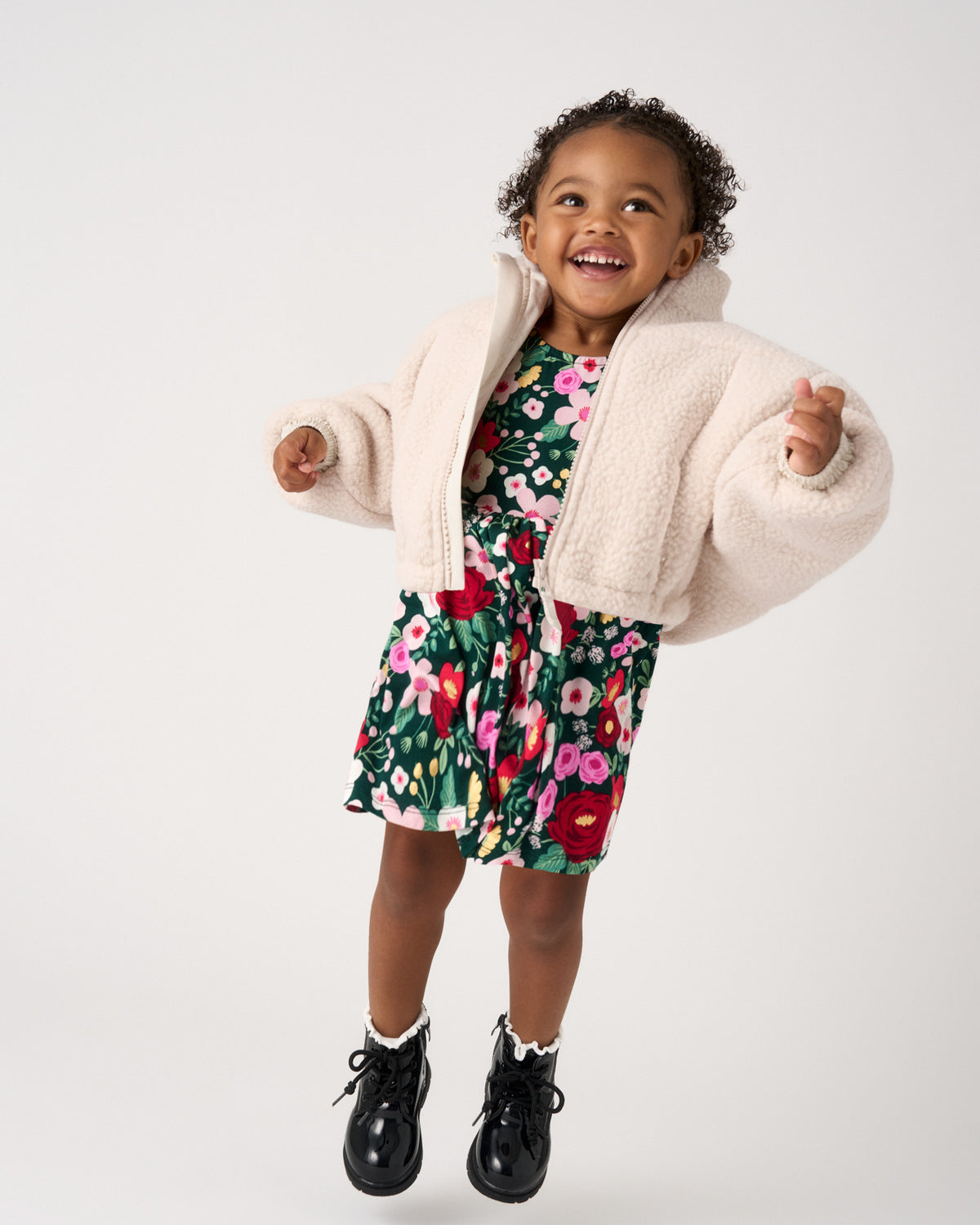 A young child wearing a cream sherpa zip-up jacket over a green floral dress, paired with black lace-up boots, captured against a solid light gray studio background.
