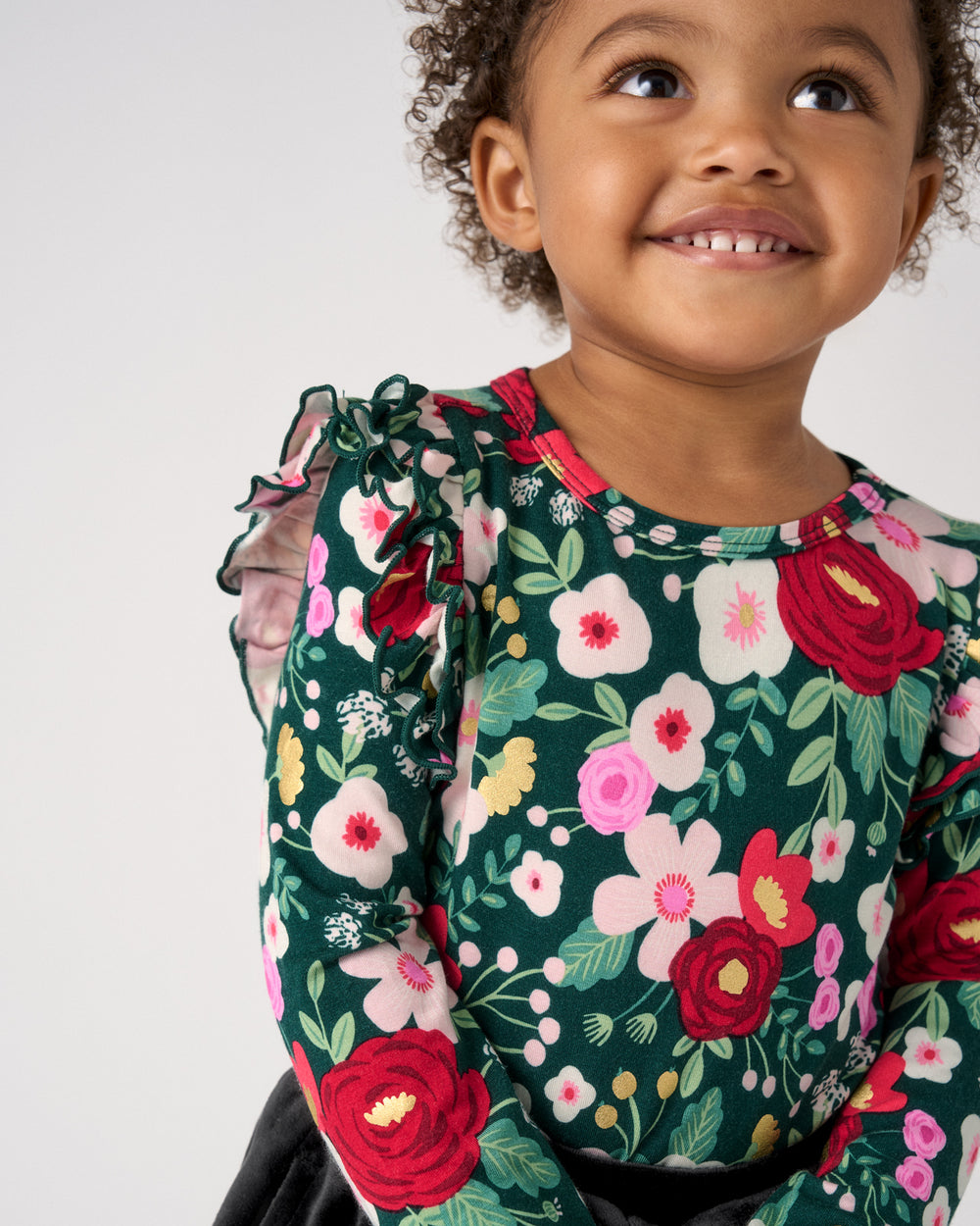 A young child wearing a dark green long-sleeve top with a vibrant floral print of red, pink, and white flowers, set against a plain light gray background.
