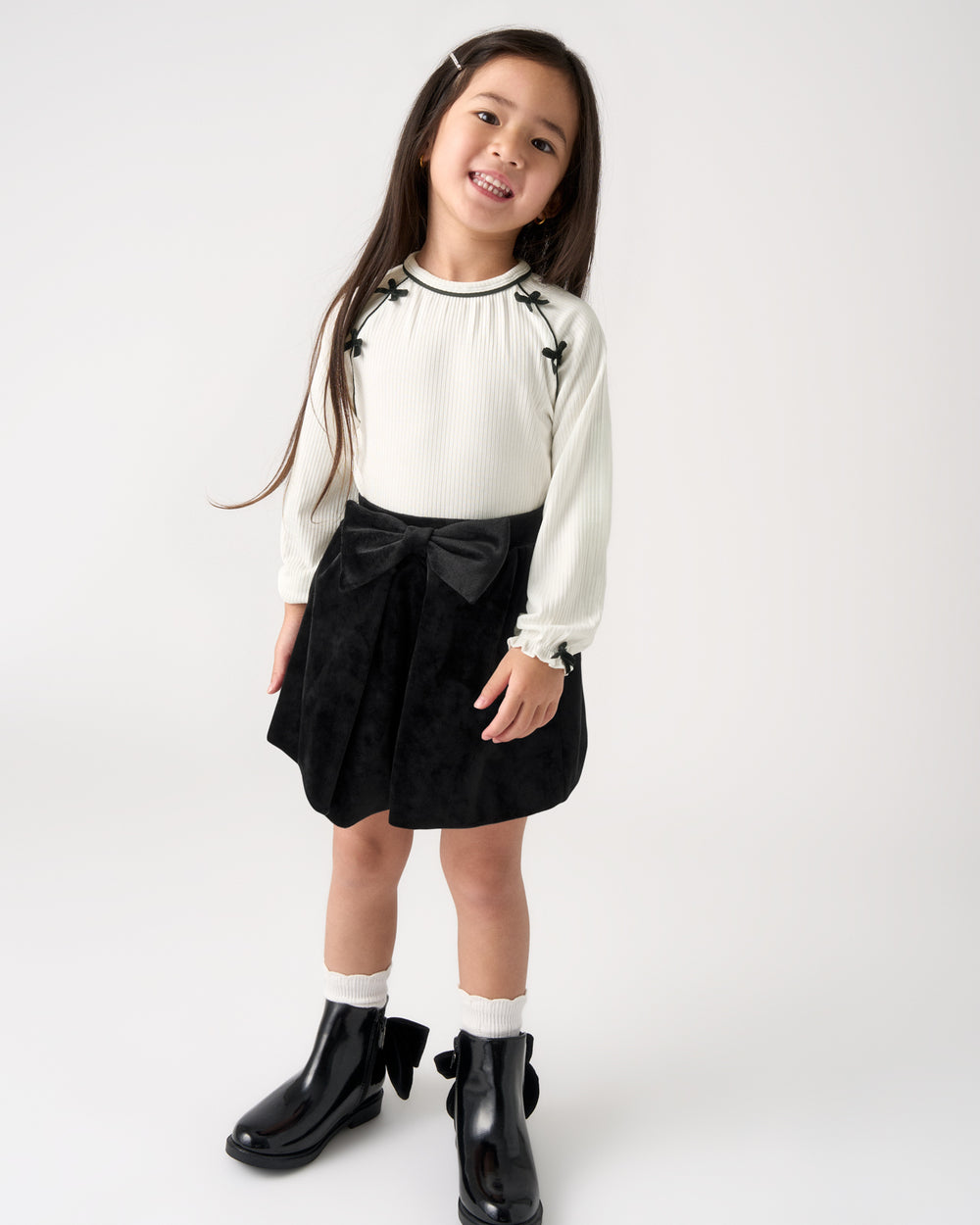 A young girl modeling a white long-sleeve blouse with black floral embroidery and a black pleated skirt featuring a large black bow at the waist, paired with white socks and black boots, against a plain white studio background.