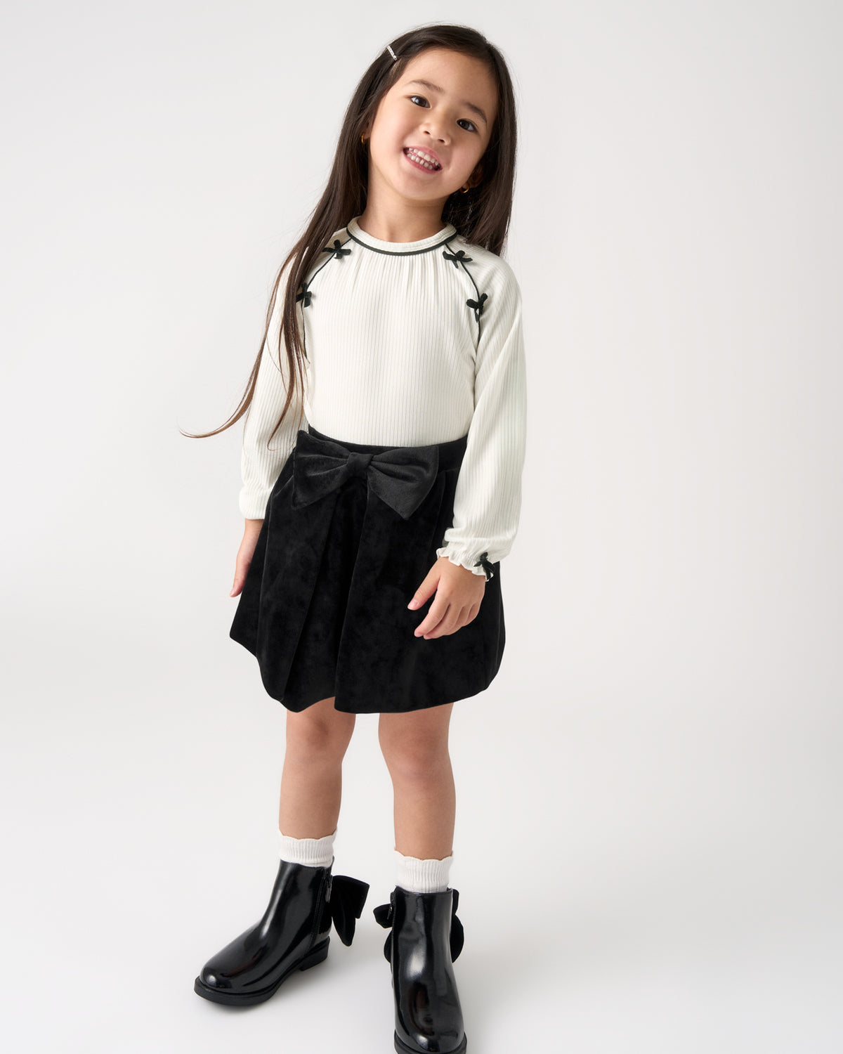 A young girl modeling a white long-sleeve blouse with black floral embroidery and a black pleated skirt featuring a large black bow at the waist, paired with white socks and black boots, against a plain white studio background.