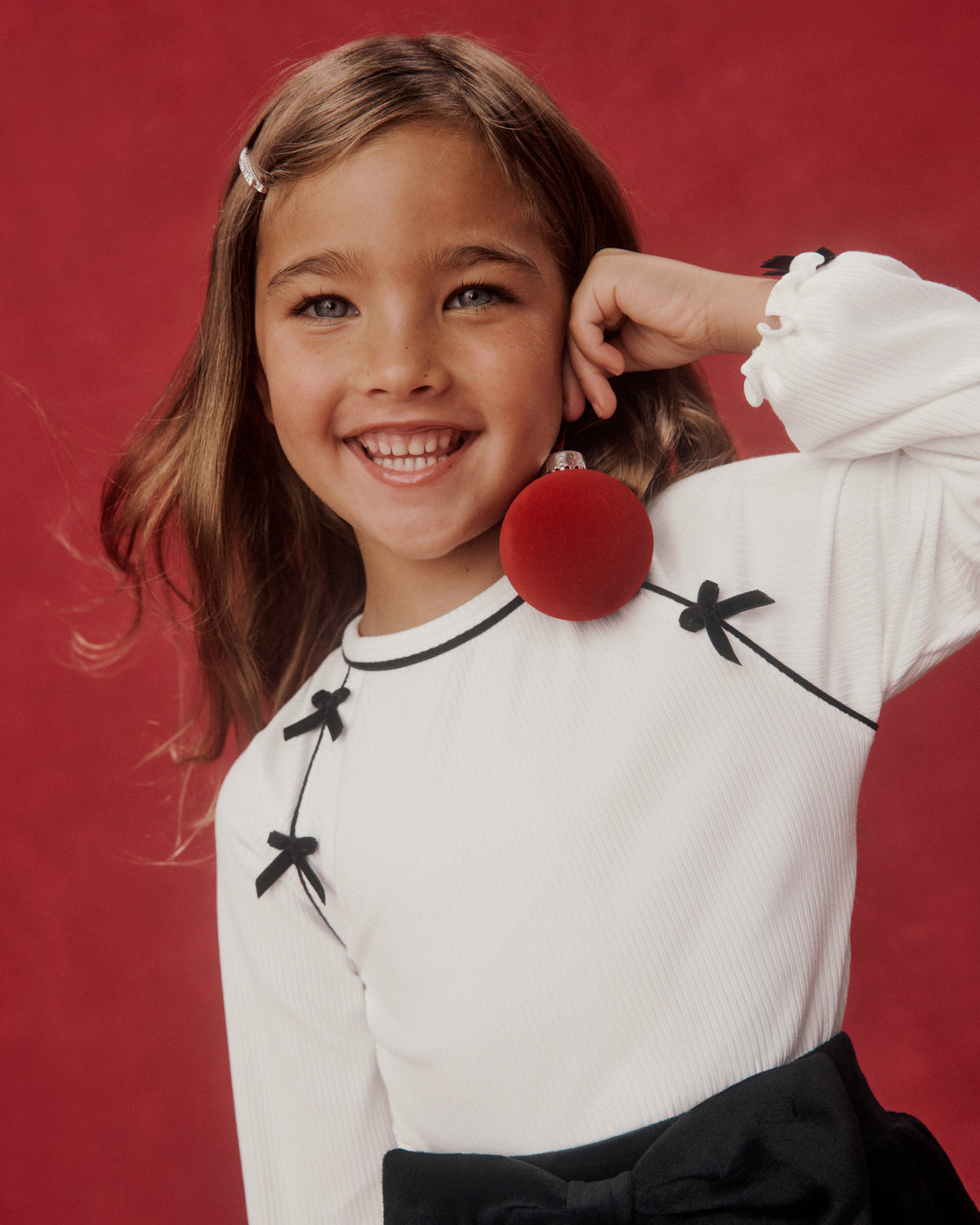 A young girl with light brown hair smiling, wearing a white ribbed long-sleeve top with black bow details and a large red pom-pom accessory, set against a solid red studio background.