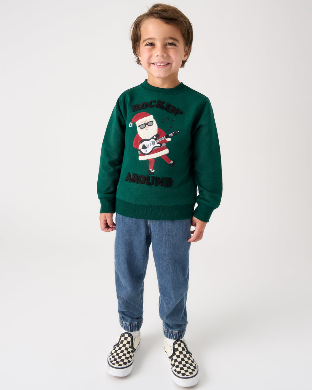 A young boy stands against a light gray studio background wearing a dark green crewneck sweatshirt with a graphic of a Santa Claus playing a guitar and the words 'ROCKIN' AROUND', paired with blue jeans and black-and-white checkered slip-on shoes.