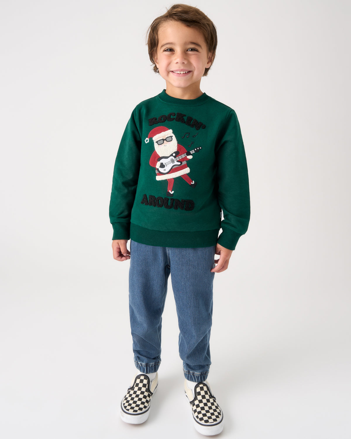 A young boy stands against a light gray studio background wearing a dark green crewneck sweatshirt with a graphic of a Santa Claus playing a guitar and the words 'ROCKIN' AROUND', paired with blue jeans and black-and-white checkered slip-on shoes.