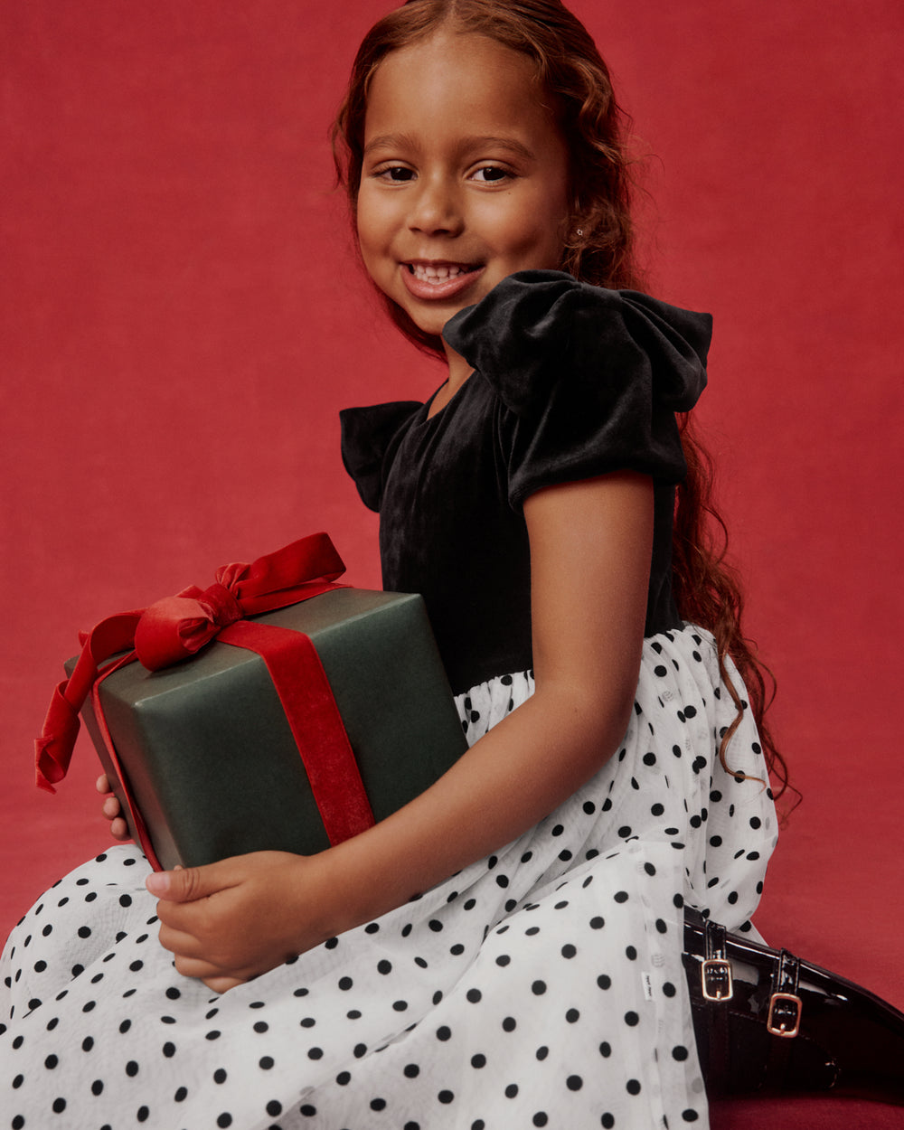 A young girl with long brown hair smiles while holding a dark green gift box with a red ribbon. She is wearing a black velvet short-sleeve top and a white dress with black polka dots, posed against a solid red studio background.