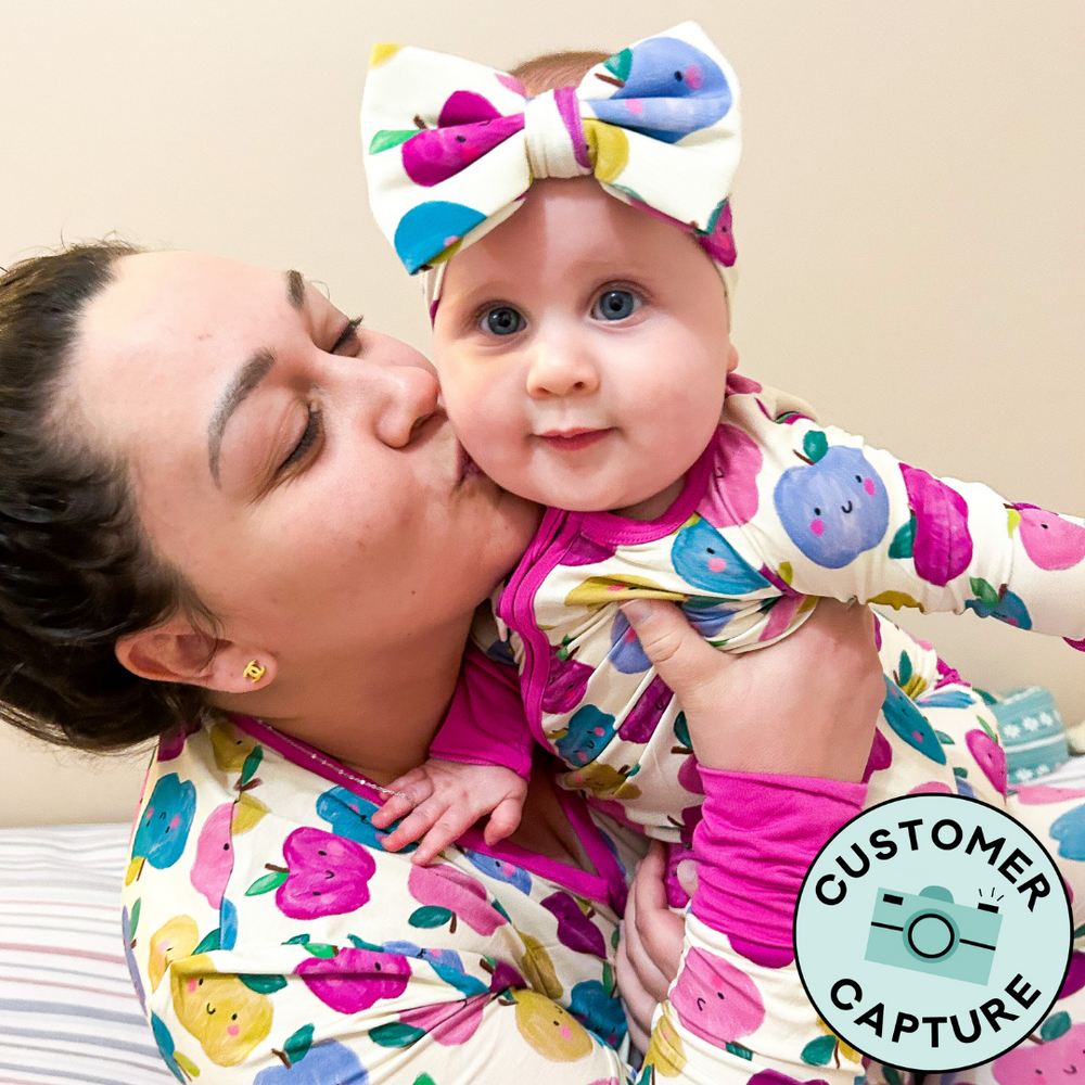 Click to see full screen - Customer Capture image of a mother and child wearing matching Berry Apple of My Eye printed pajamas. Mom is wearing a Berry Apple of My Eye women's pajama top and her child is matching wearing a zippy paired with a luxe bow headband.