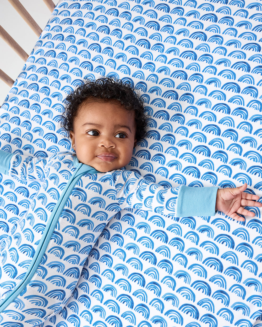 Baby wearing the Blue Rainbows Fitted Crib Sheet and coordinating zippy