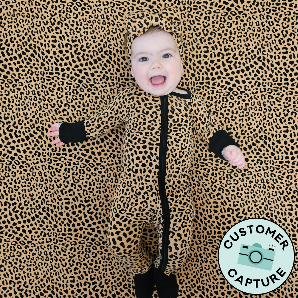 Customer Capture image of a child laying on a Classic Leopard crib sheet wearing a matching Classic Leopard zippy paired with a matching luxe bow headband