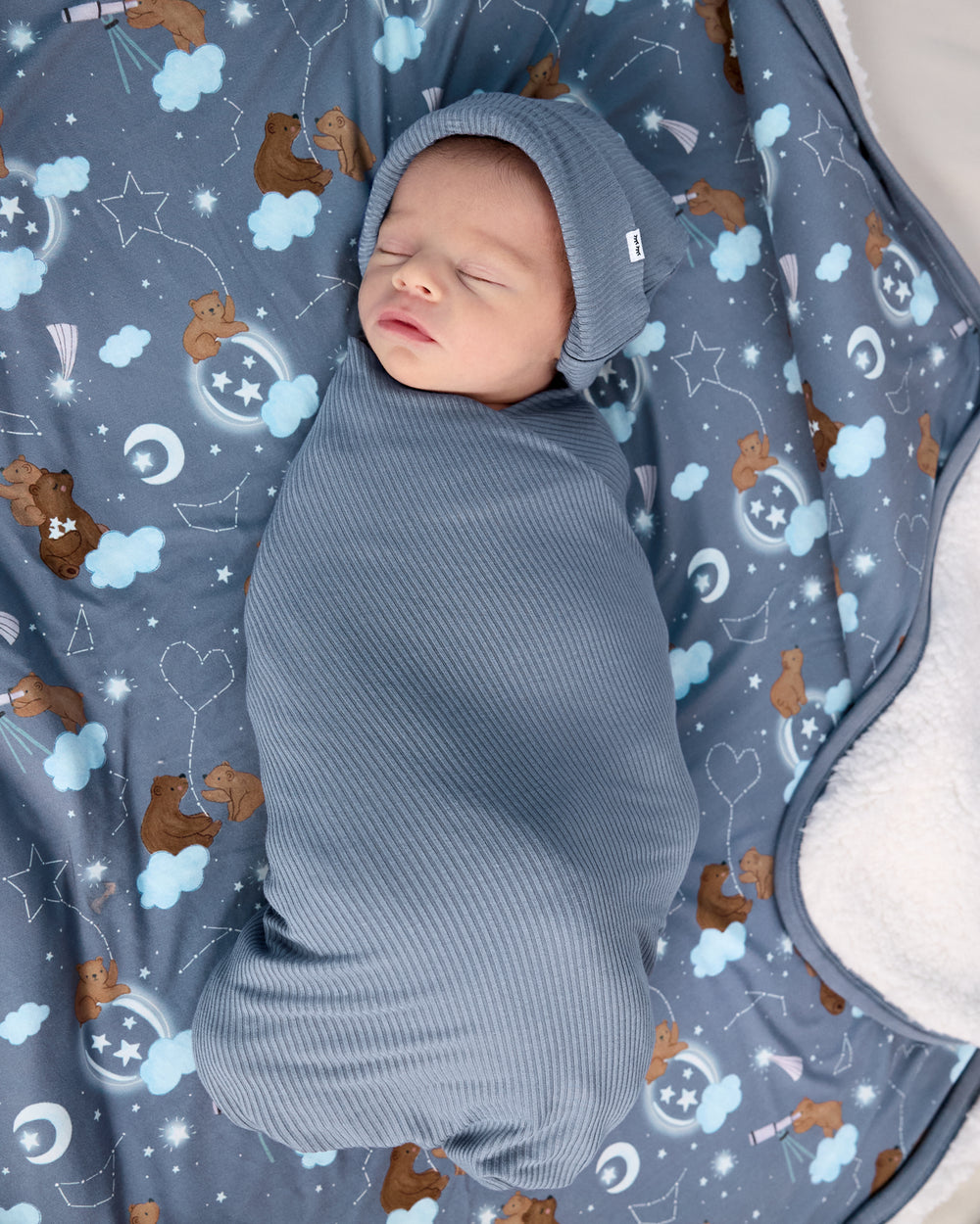 Newborn wearing the Hazy Blue Ribbed Swaddle & Infant Beanie Set on the Constellation Cubs sherpa blanket
