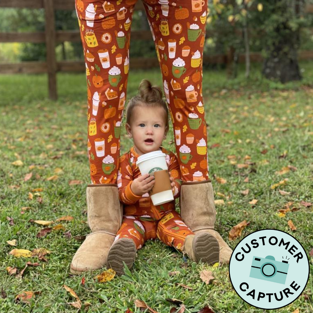 Customer Capture image of a child sitting between her mother's legs. Mom is wearing Pumpkin Spice women's pajama pants and child is wearing a Pumpkin Spice zippy