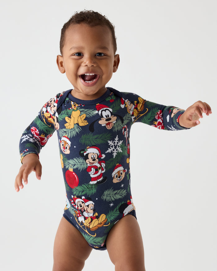 A smiling baby wearing a long-sleeve navy blue bodysuit covered in a colorful Mickey Mouse and Christmas holiday print, posed against a light gray studio background.