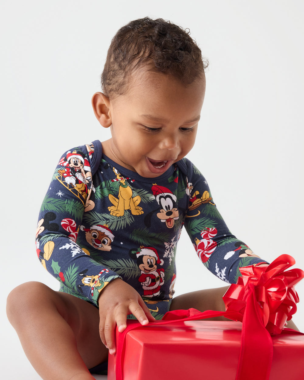 A toddler wearing a navy blue long-sleeve pajama top with a colorful Disney character holiday print, sitting and reaching for a large red gift box with a red ribbon bow against a light gray background.