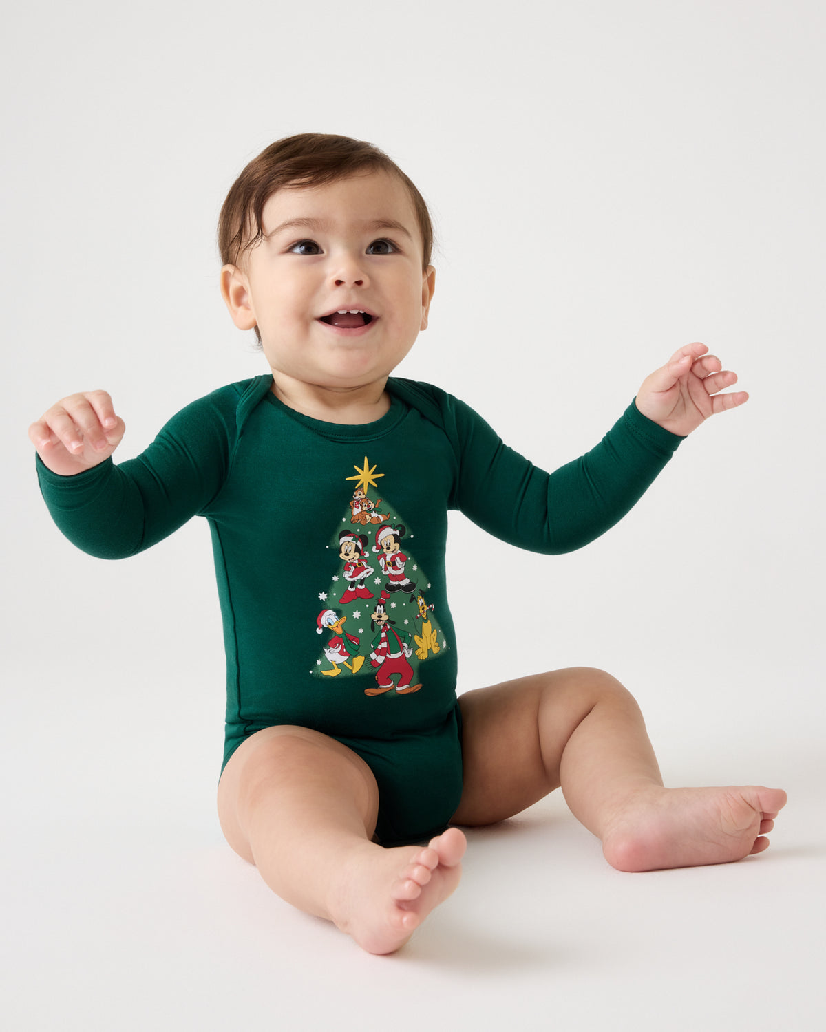 A baby sitting on a white studio floor wearing a dark green long-sleeve bodysuit with a colorful Christmas tree graphic on the front.