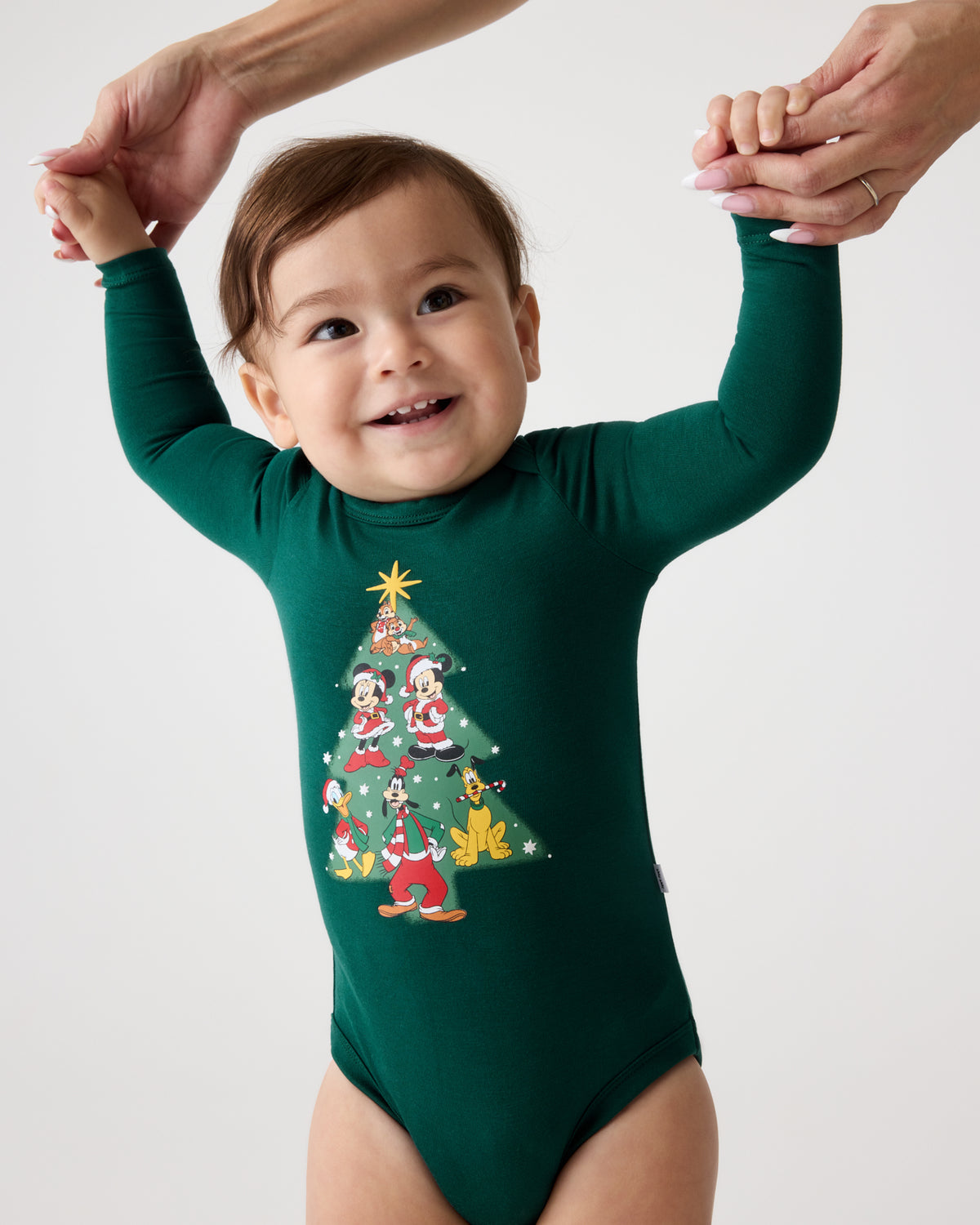 A baby wearing a dark green long-sleeve bodysuit with a colorful Christmas tree graphic on the front, holding hands with an adult against a plain light background.