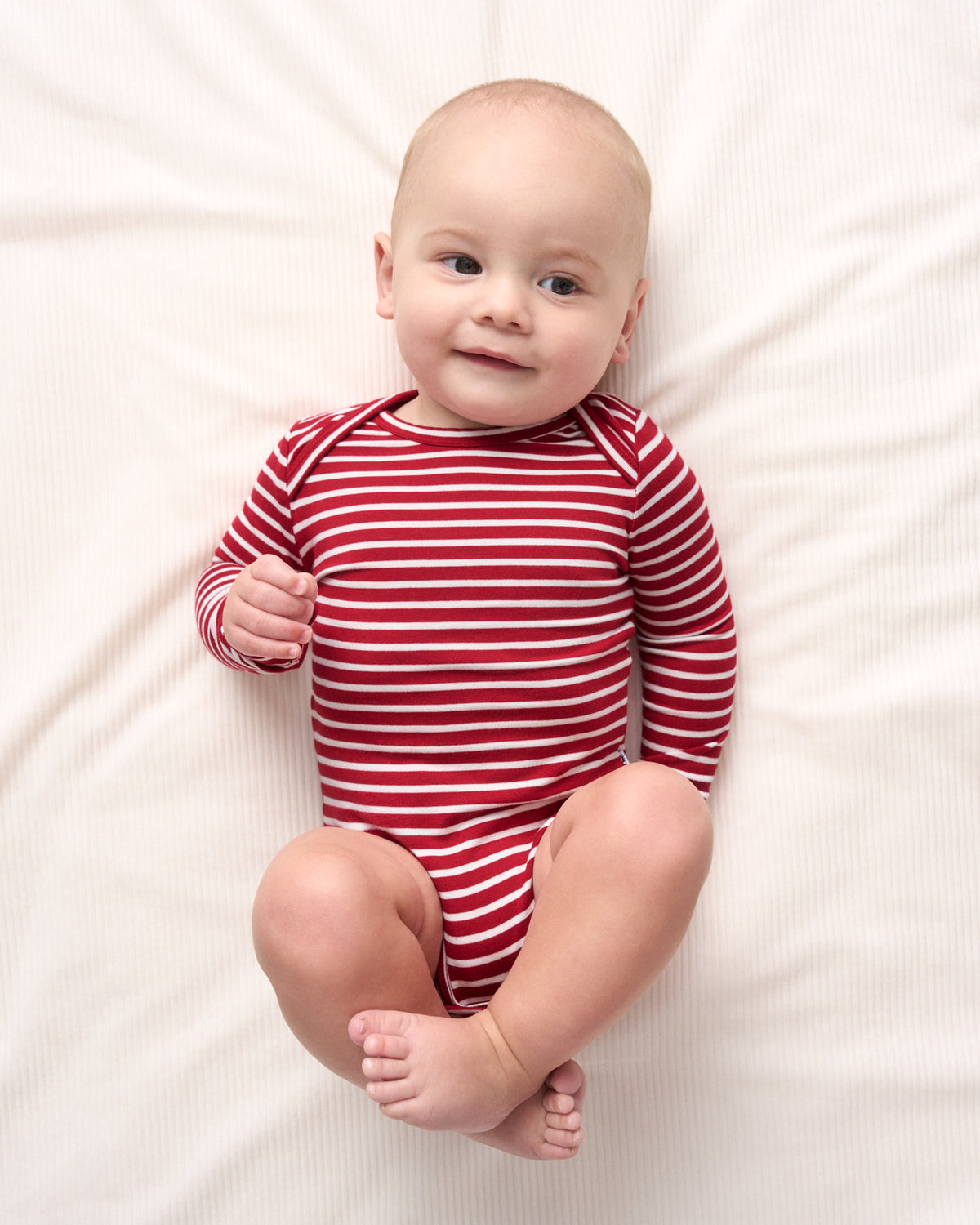 A baby lying on a white textured blanket, wearing a red and white horizontally striped long-sleeve bodysuit.