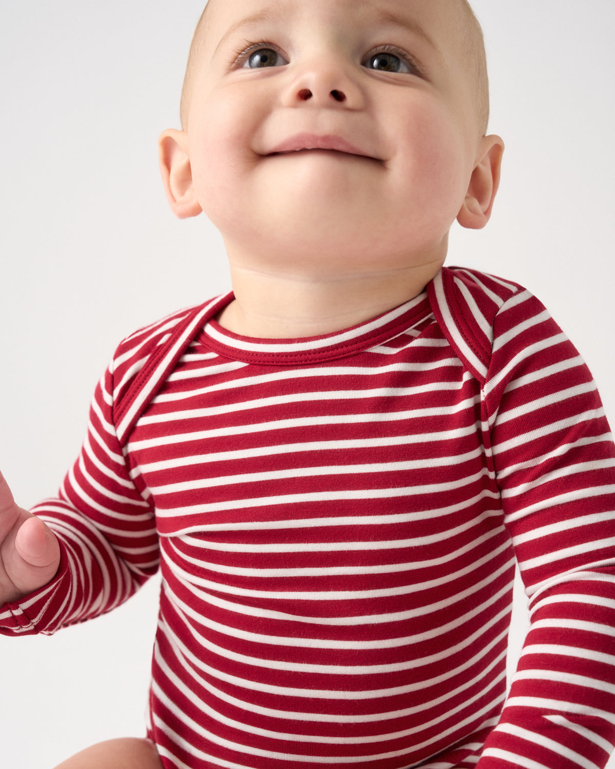 A close-up of a smiling baby wearing a red and white horizontally striped long-sleeve bodysuit, photographed against a clean light gray background.