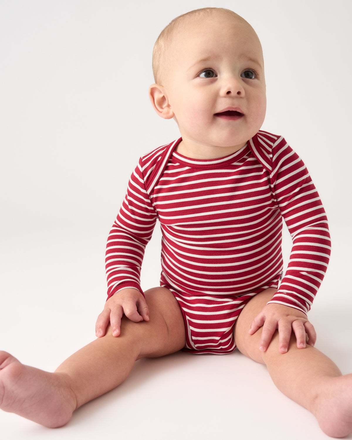 A baby sitting on a white studio background wearing a red and white horizontally striped long-sleeve bodysuit.