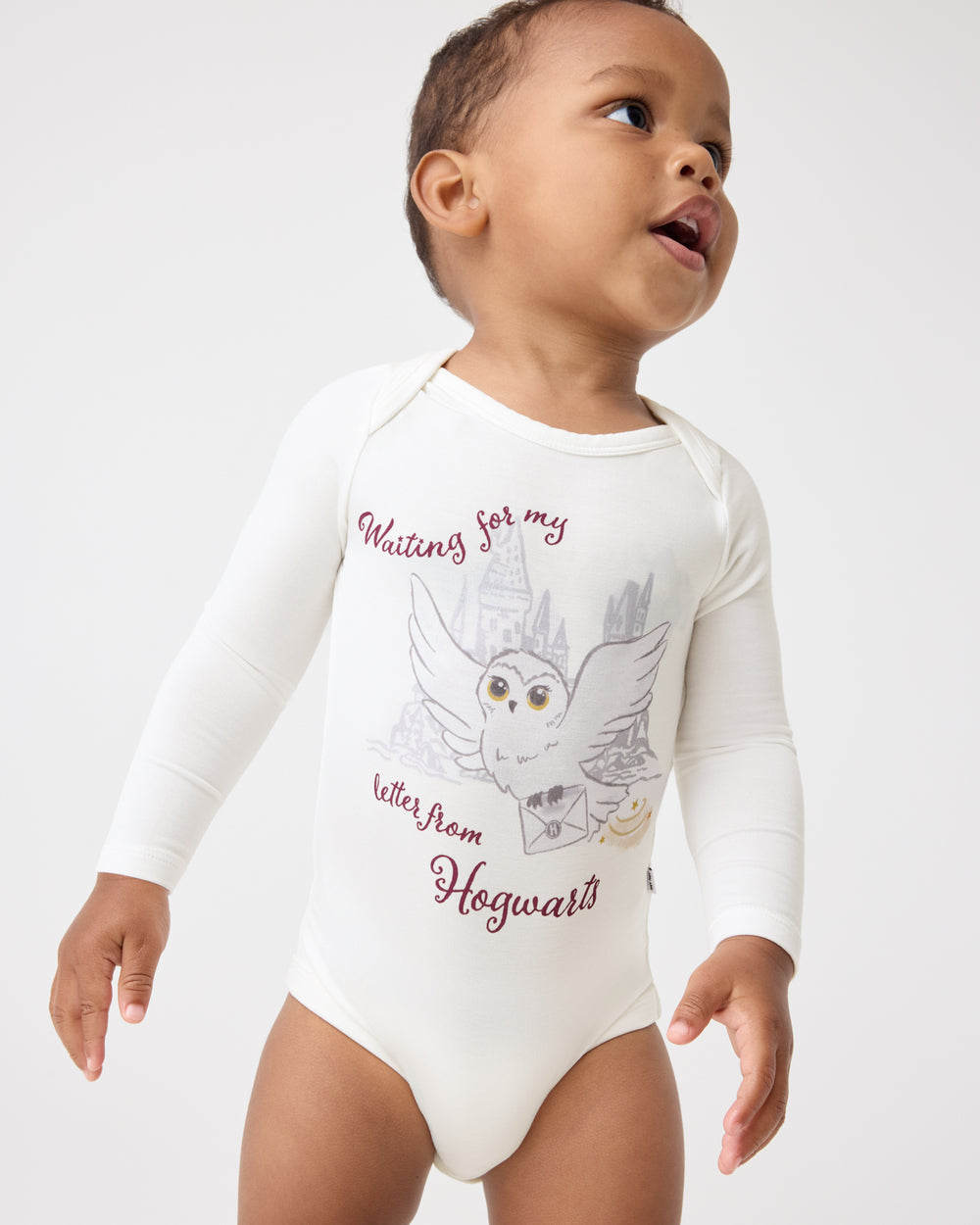 A baby wearing a Harry Potter™ Hedwig Bodysuit featuring white long-sleeves with a Harry Potter owl and castle graphic and the text 'Waiting for my letter from Hogwarts' on a light gray background.