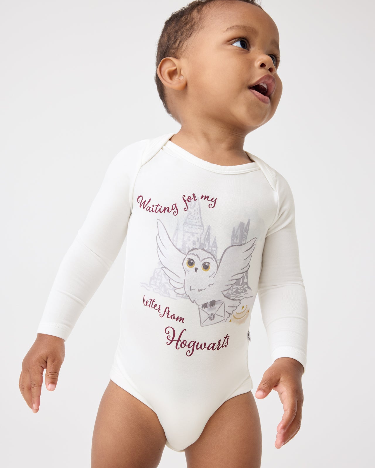 A baby wearing a Harry Potter™ Hedwig Bodysuit featuring white long-sleeves with a Harry Potter owl and castle graphic and the text 'Waiting for my letter from Hogwarts' on a light gray background.