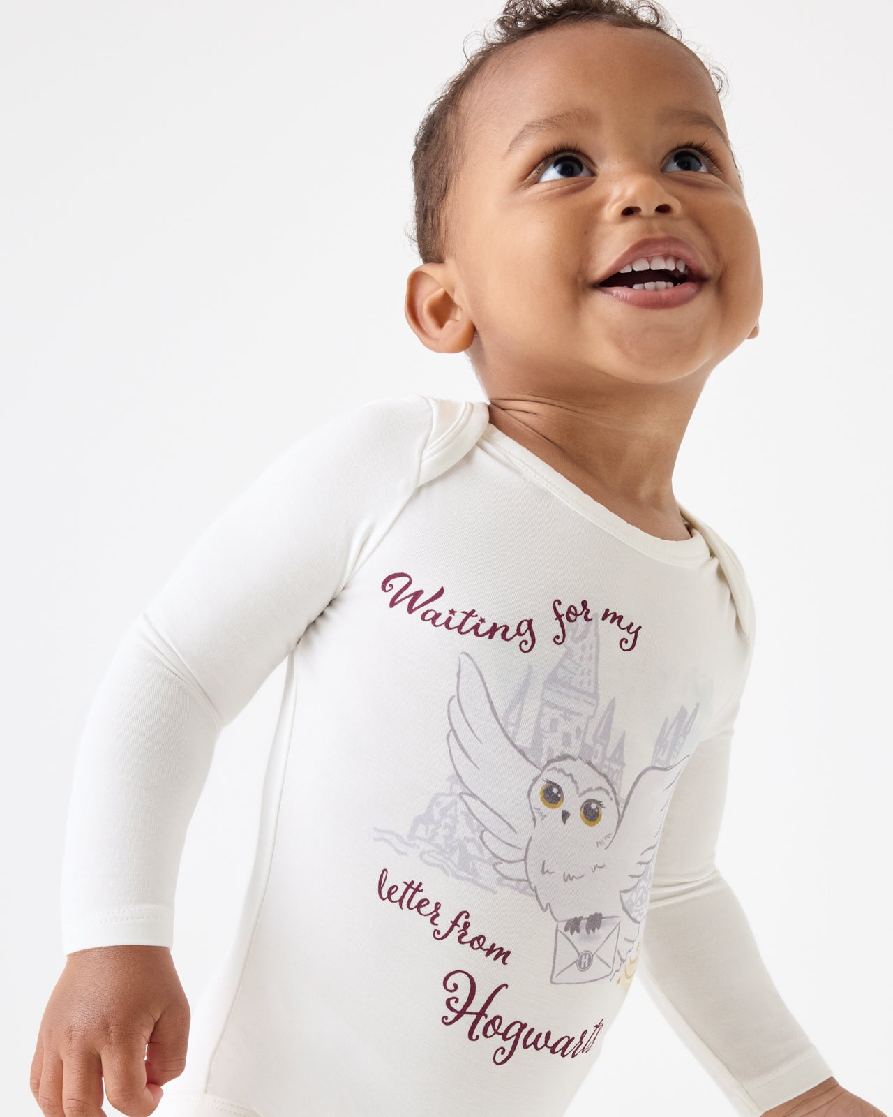 Close up of baby wearing a Harry Potter™ Hedwig Bodysuit featuring white long-sleeves with a Harry Potter owl and castle graphic and the text 'Waiting for my letter from Hogwarts' on a light gray background.