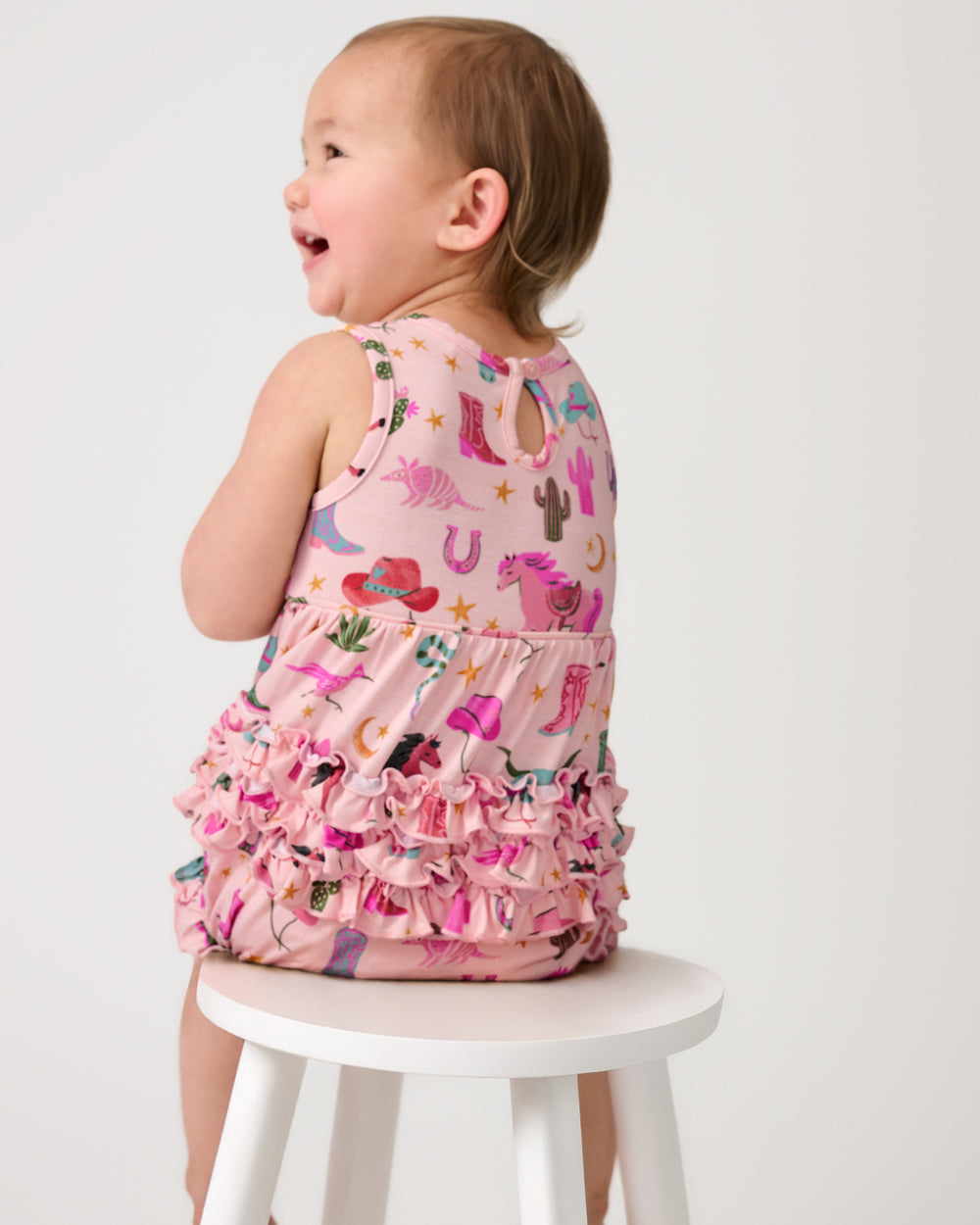Back view of child sitting on a white bench wearing the Pink Rancher Roundup Bubble Romper
