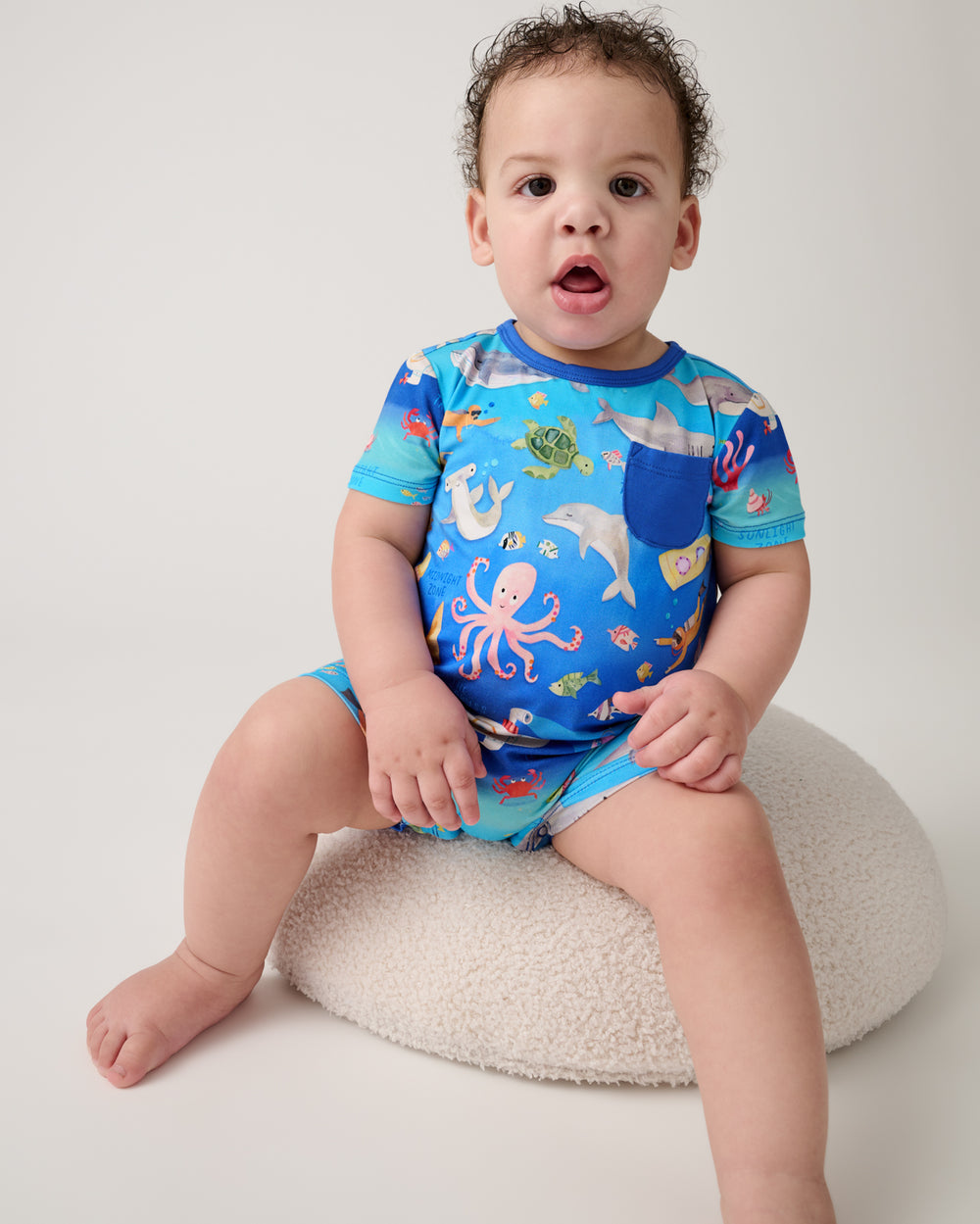 Baby sitting on a white ottoman wearing the Underwater Discovery Pocket Shorty Romper