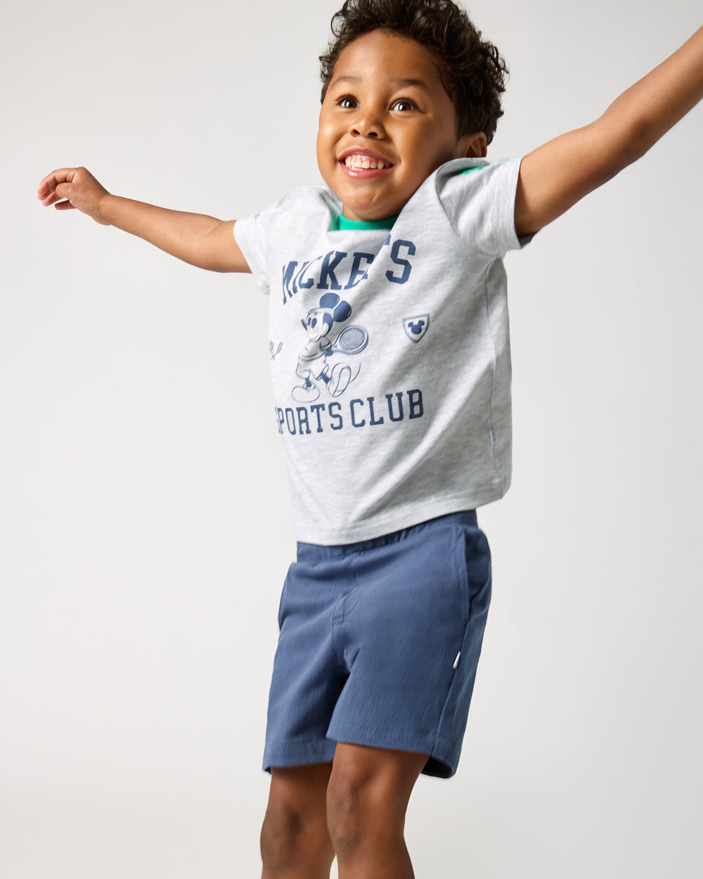 Jumping boy wearing the Disney Mickey Mouse Sports Club Relaxed Tee with navy shorts