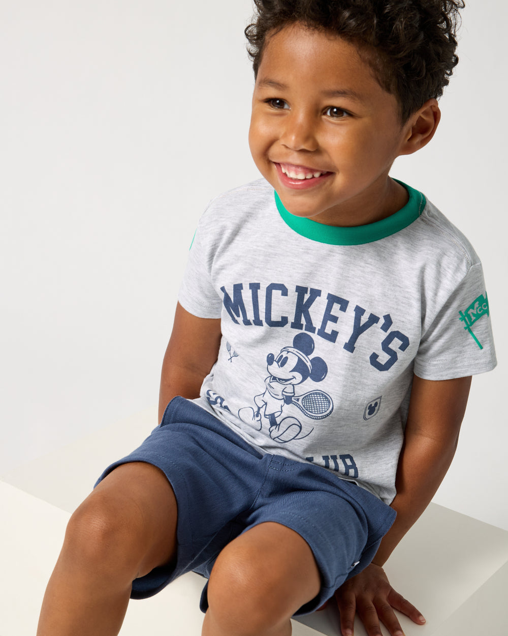 Sitting boy wearing the Disney Mickey Mouse Sports Club Relaxed Tee with navy shorts