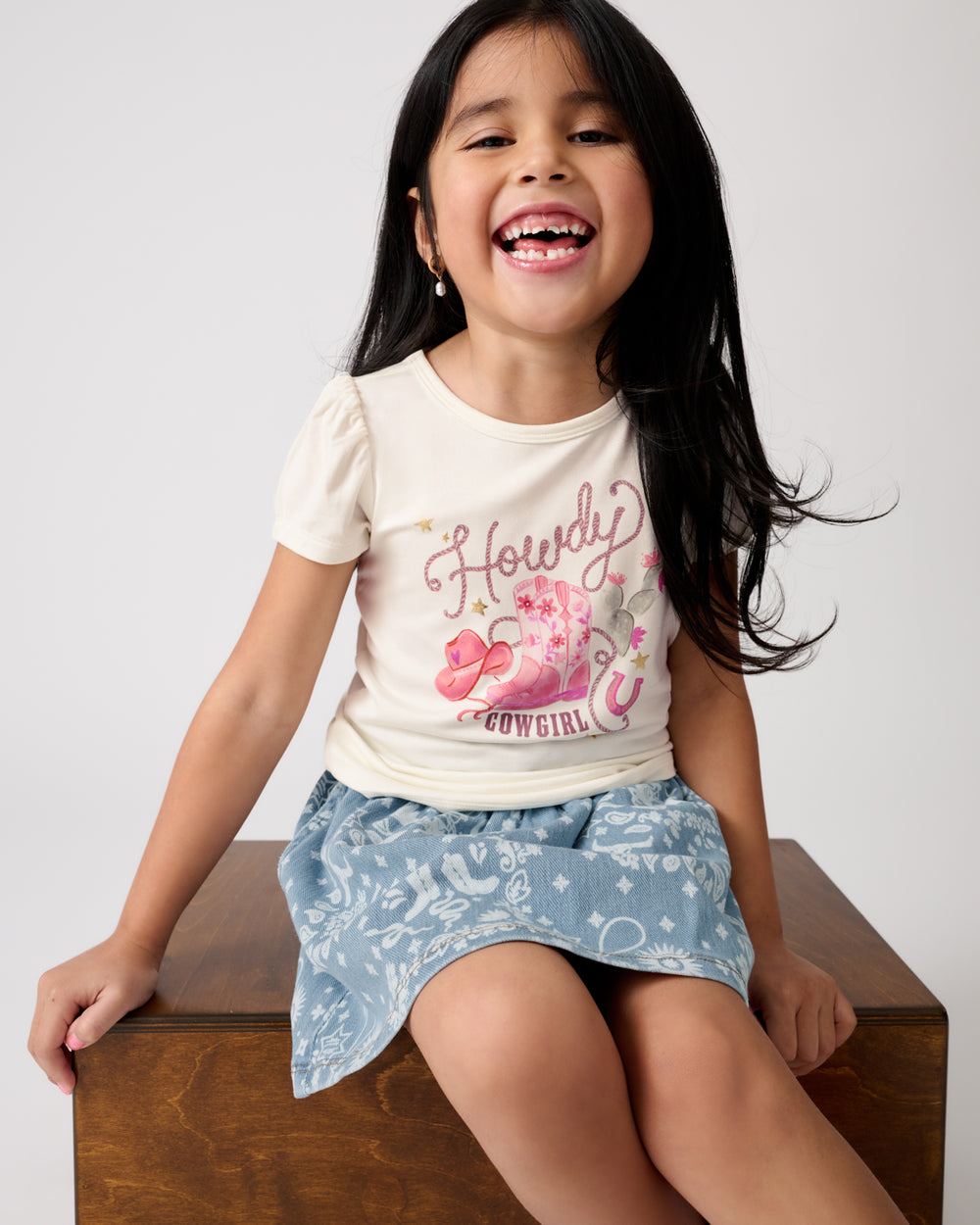 Girl sitting on a wooden box wearing the Howdy Cowgirl Puff Sleeve Tee and Bandana Print Skort