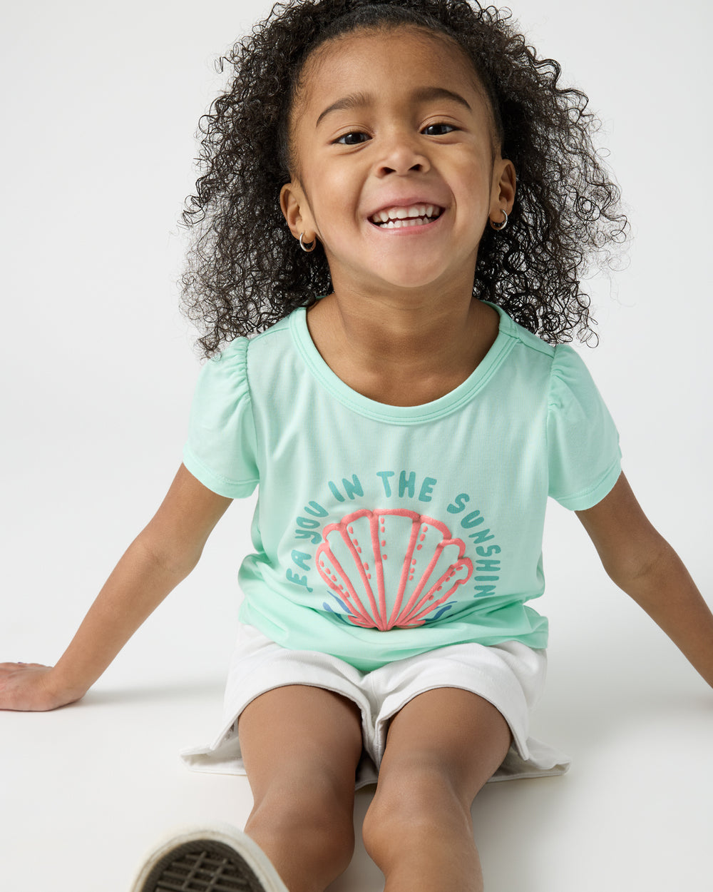 Sitting girl wearing the Sparkling Seas Puff Sleeve Tee with white shorts
