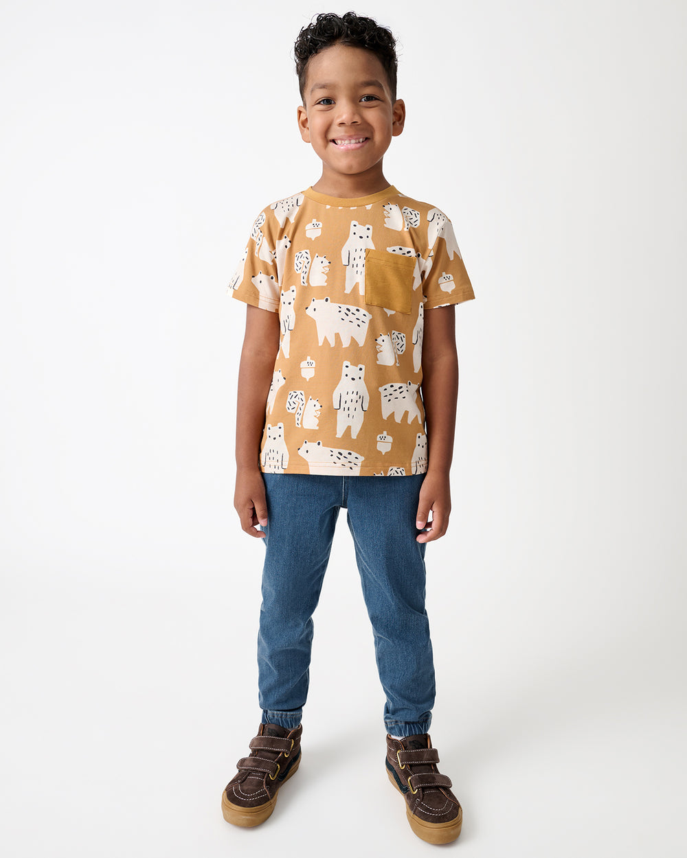 A young boy stands against a light gray studio background wearing a mustard yellow short-sleeve t-shirt with a white animal print, blue jeans, and brown shoes.