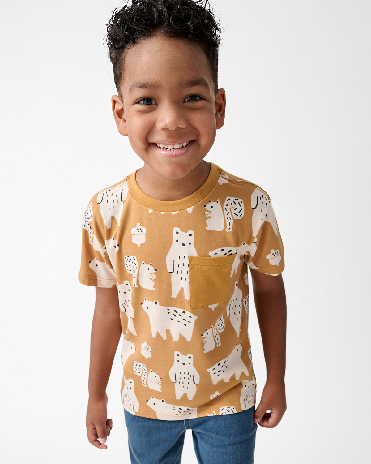 A smiling young boy wearing a mustard yellow short-sleeve t-shirt with a white bear pattern, paired with blue jeans, photographed against a clean white background.