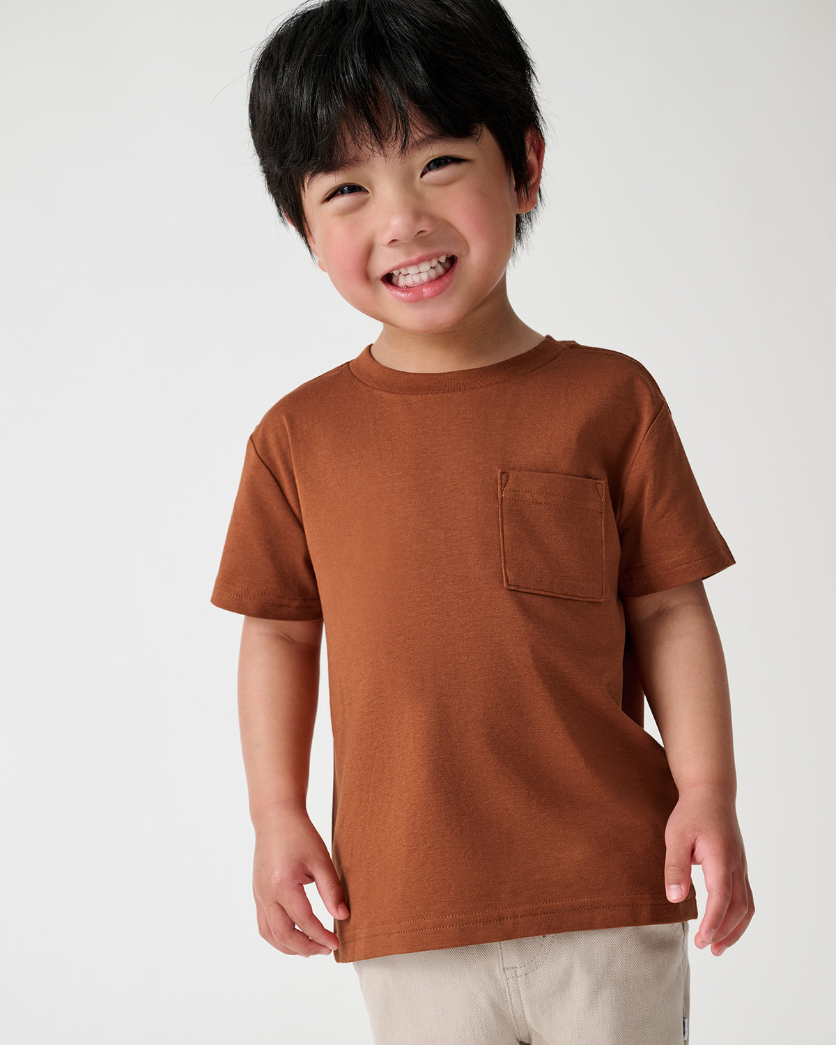 A smiling young boy wearing a rust-brown short-sleeve crewneck t-shirt with a chest pocket, paired with light beige pants, photographed against a clean white studio background.