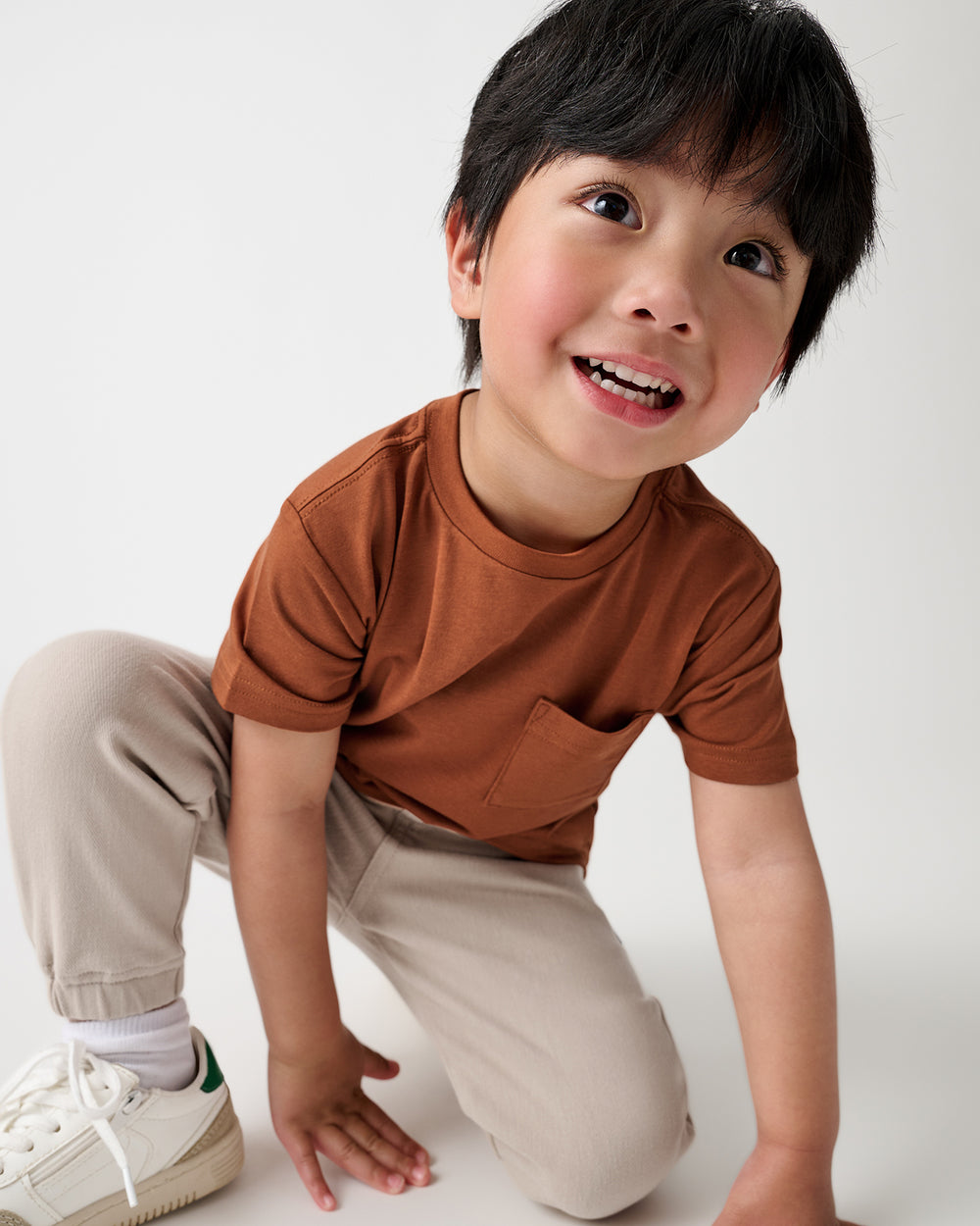 A young boy wearing a rust-brown short-sleeve pocket t-shirt, beige pants, and white sneakers, kneeling down in a playful pose against a clean white studio background.