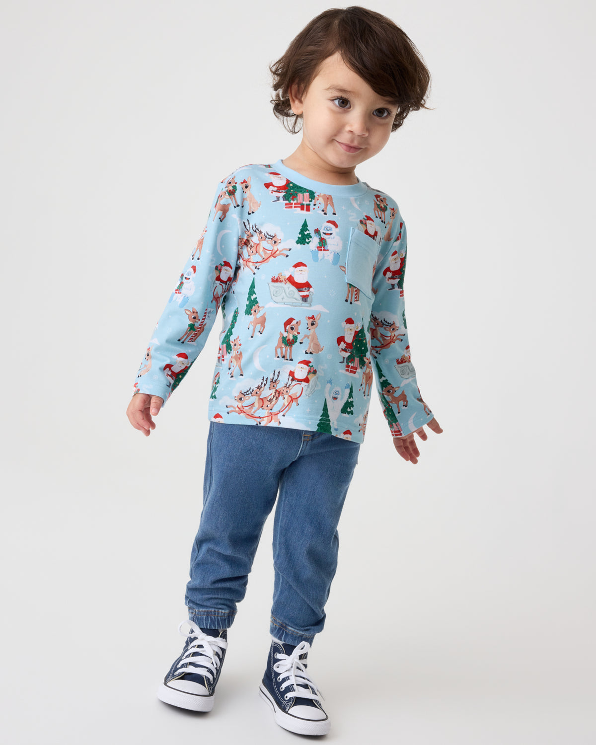A toddler standing against a light gray studio background, wearing a light blue long-sleeve shirt with a festive Christmas print of Santa Claus, reindeer, trees, and snowflakes, paired with blue jeans and navy sneakers.