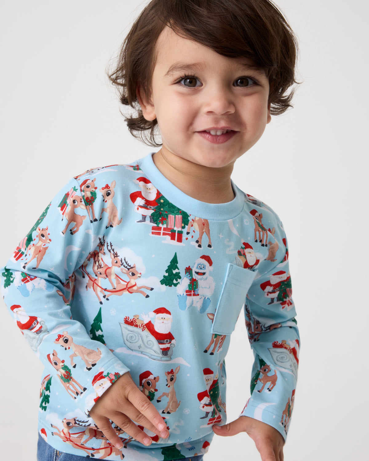 A smiling young child wearing a light blue long-sleeve pajama top with a festive Christmas print of Santa Claus, reindeer, and snow trees, photographed against a plain light gray background.