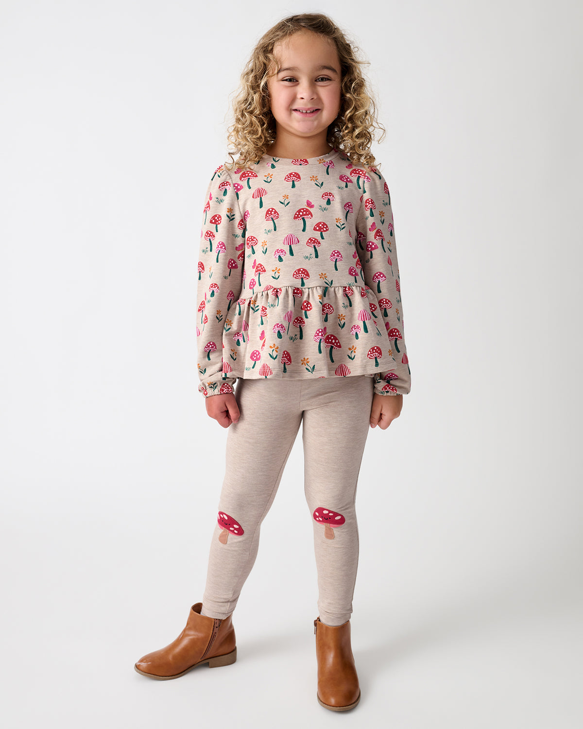 A young girl stands against a light gray studio background wearing a beige long-sleeve top with a colorful mushroom print, matching beige leggings with red mushroom patches on the knees, and brown ankle boots.