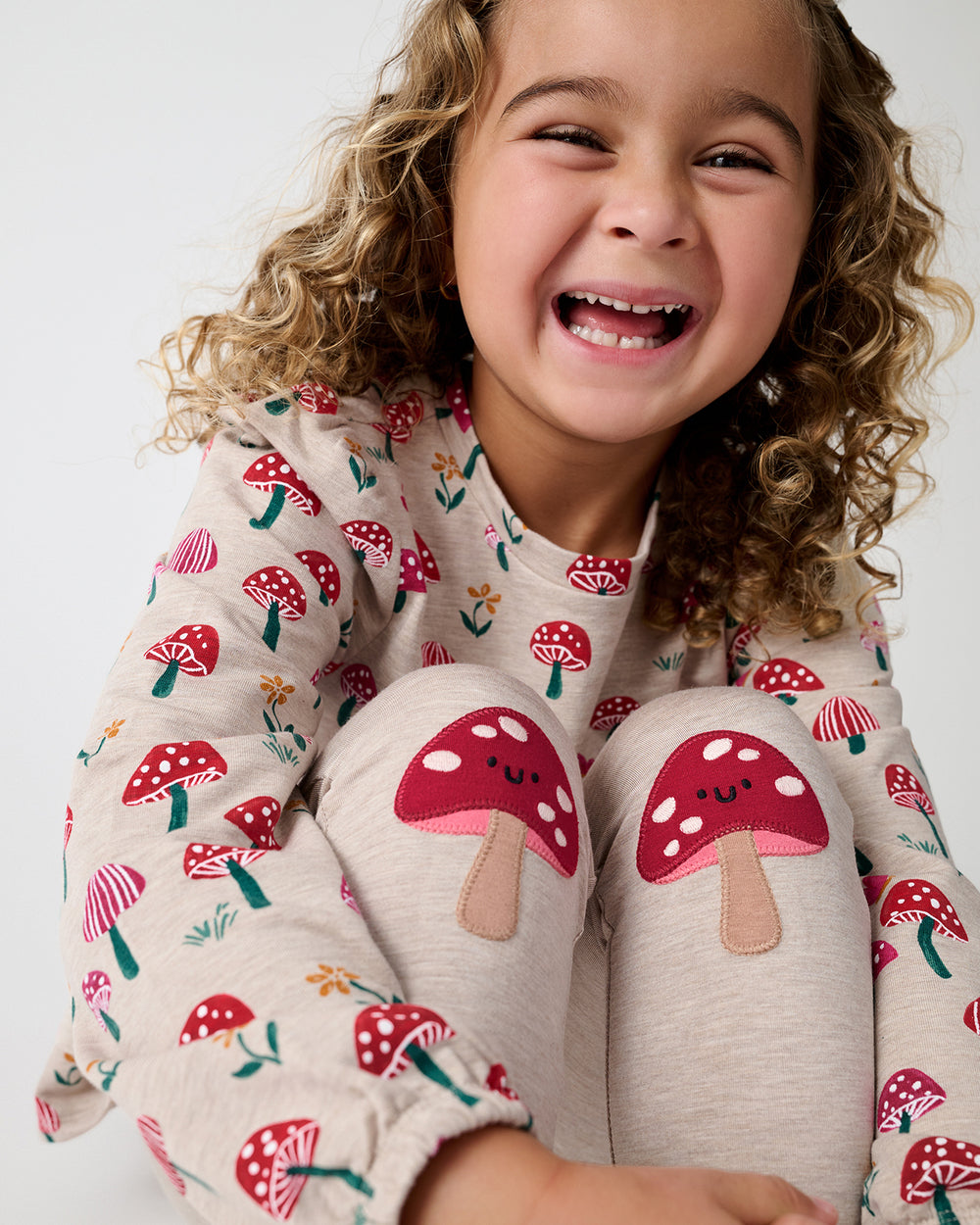 A smiling young girl wearing a cozy long-sleeve pajama top and matching pants with a red and white mushroom print, sitting against a plain light background.