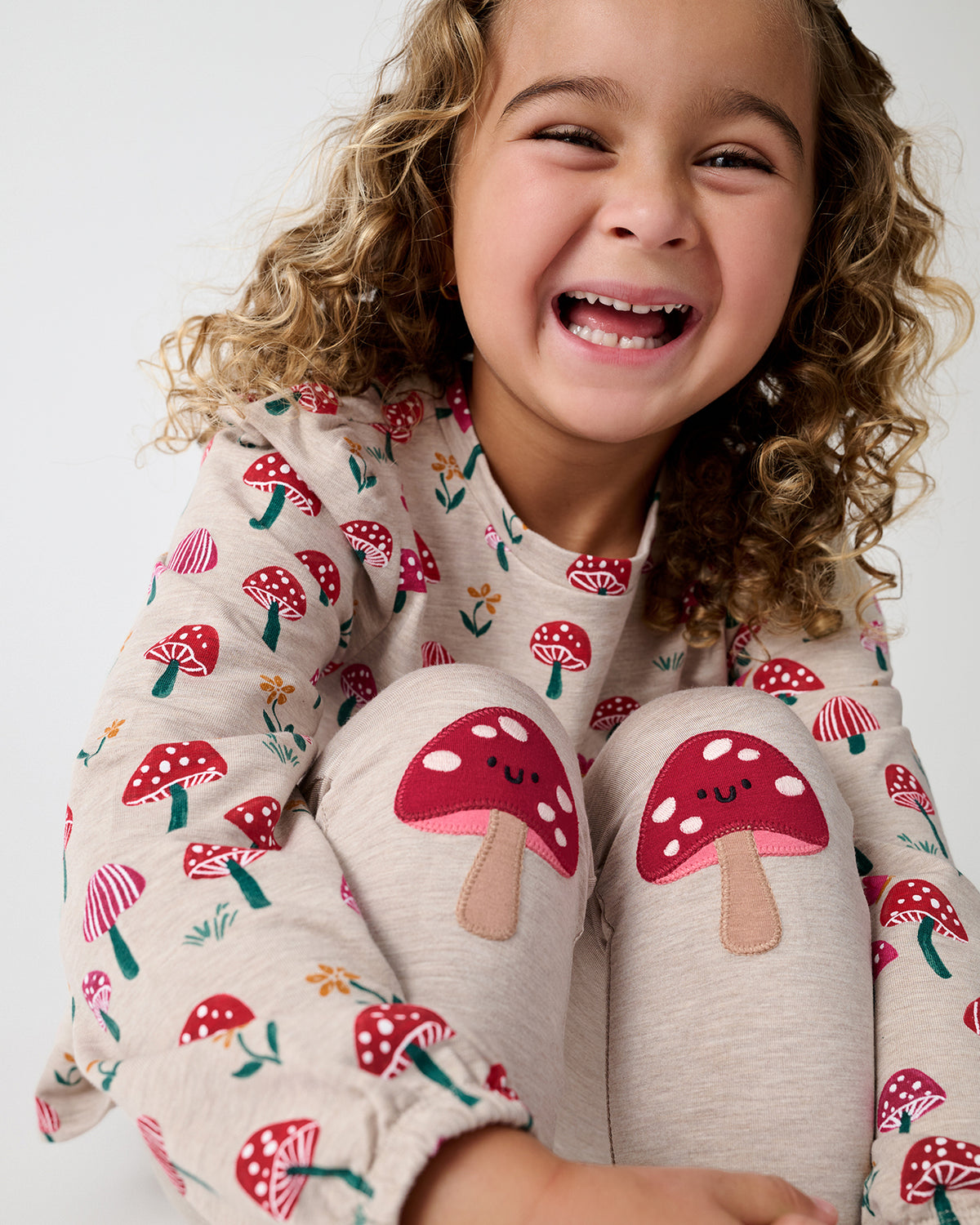 A smiling young girl wearing a cozy long-sleeve pajama top and matching pants with a red and white mushroom print, sitting against a plain light background.