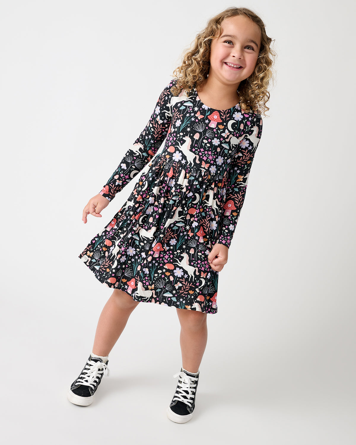 A young girl smiling and posing in a long-sleeve black dress with a colorful woodland animal and floral pattern, paired with black and white sneakers, against a plain light gray studio background.
