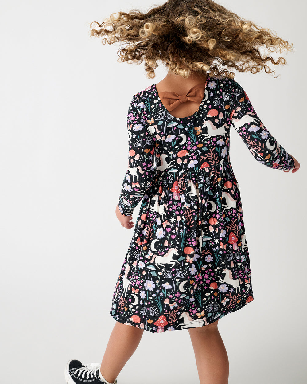 A young girl is shown from a back angle wearing a long-sleeve dress with a dense multicolor whimsical, floral and butterfly print on a black background. She is wearing black and white sneakers against a clean white studio background.