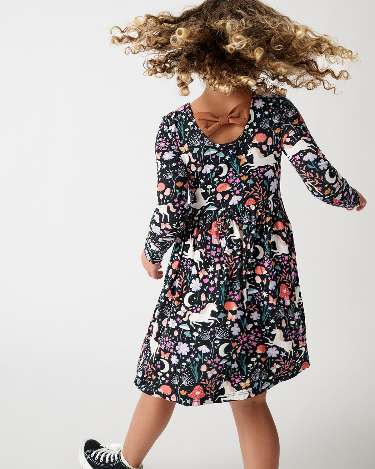 A young girl is shown from a back angle wearing a long-sleeve dress with a dense multicolor whimsical, floral and butterfly print on a black background. She is wearing black and white sneakers against a clean white studio background.