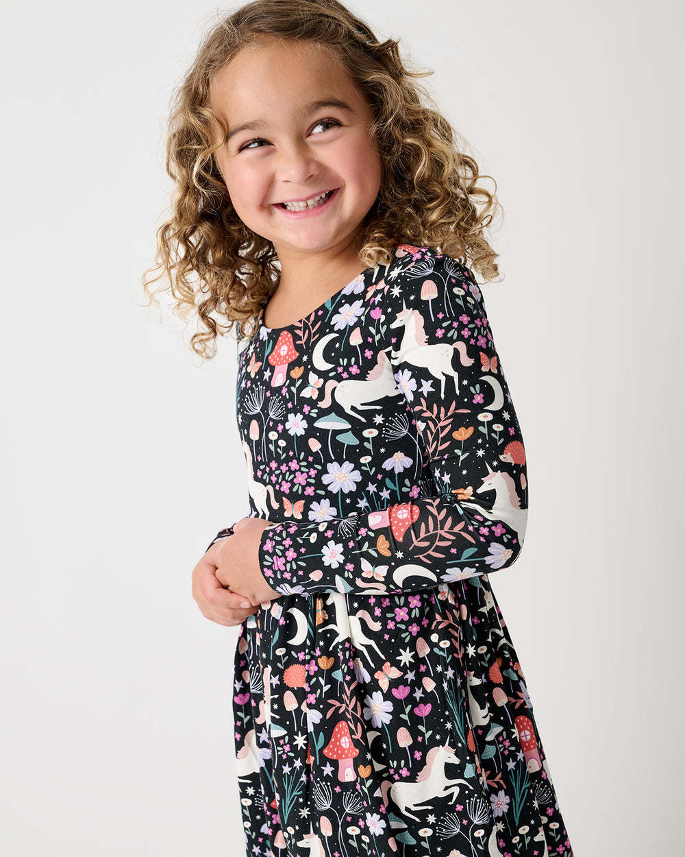 A young girl smiling and looking to the side, wearing a long-sleeve black dress with a colorful woodland print of white unicorns, pink mushrooms, and green leaves, set against a plain light gray background.