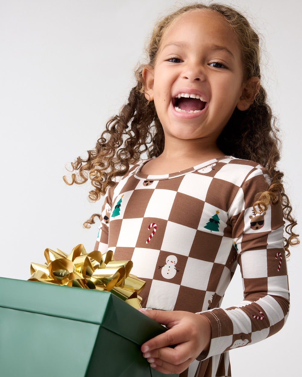 A young girl smiles while holding a green gift box with a large gold bow. She is wearing a long-sleeve dress with a brown and white checkerboard pattern decorated with small holiday motifs like candy canes, a snowman, and a Christmas tree, against a plain light gray studio background.