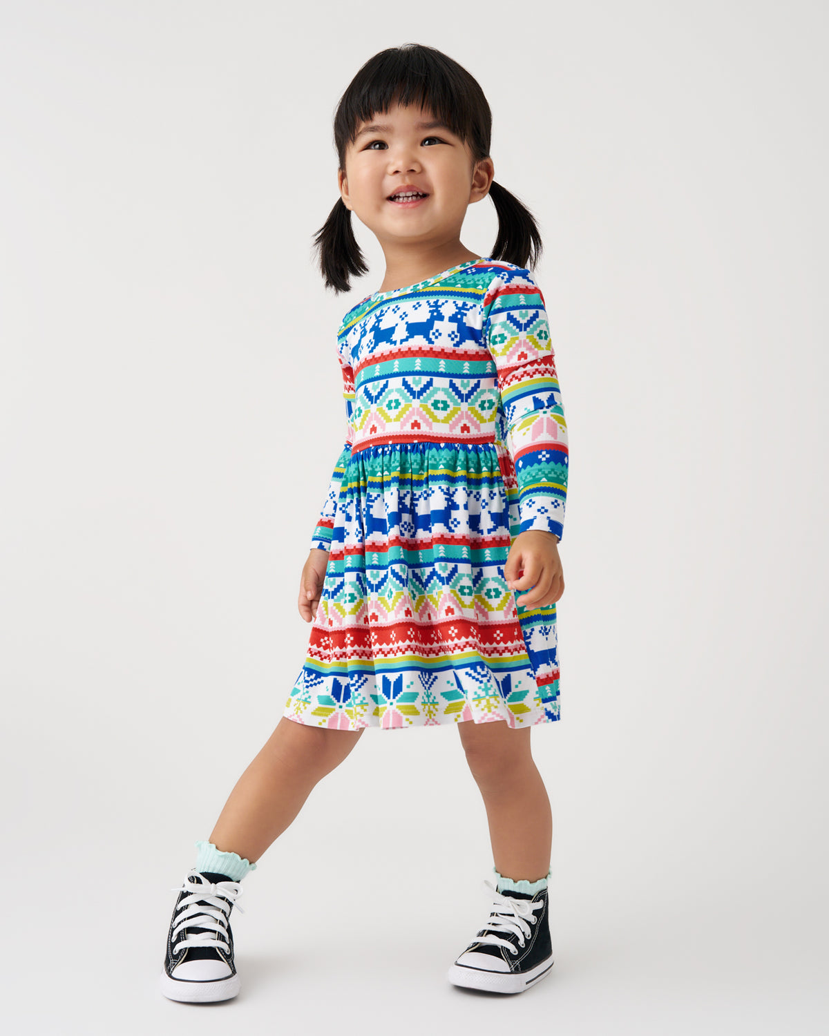 A young girl with dark hair in pigtails stands against a light gray studio background wearing a long-sleeve tiered dress with a colorful geometric pattern in blue, red, yellow, green, and white. She is also wearing black and white high-top sneakers.