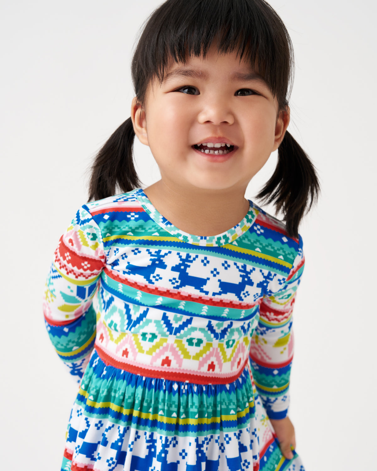A young girl with dark hair in pigtails smiling, wearing a long-sleeve knit dress with a colorful Nordic fair isle pattern featuring blue, green, red, yellow, and white.