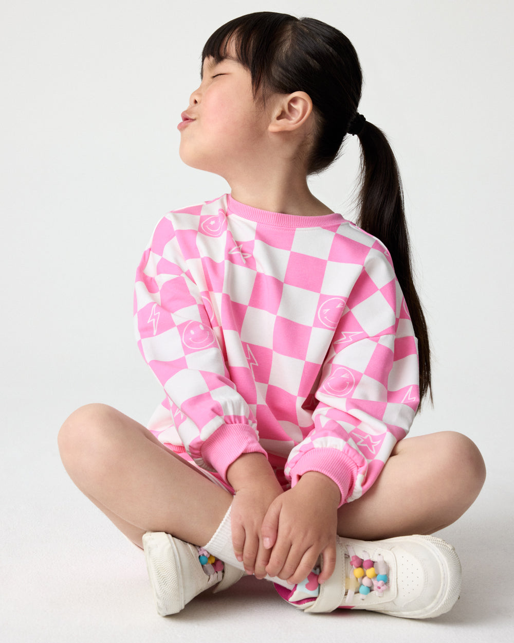 Sitting child wearing the Cheery Checks Pink Drop Shoulder Sweatshirt with white sneakers