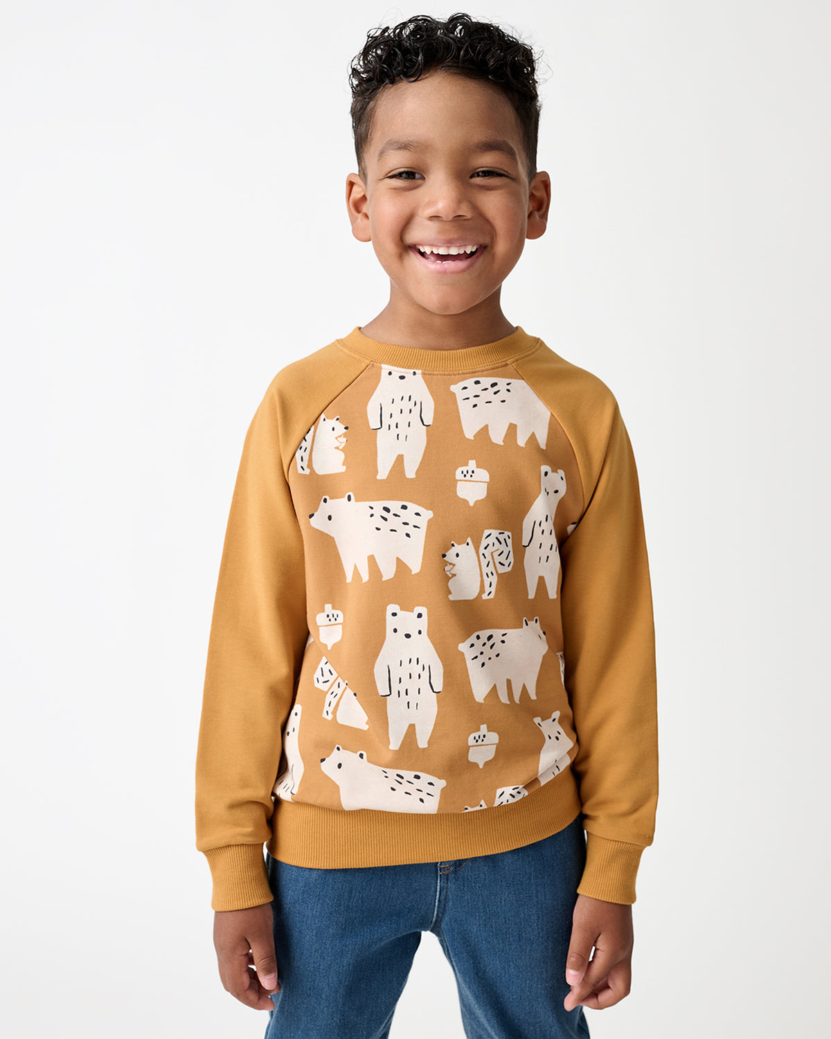 A smiling young boy wearing a mustard yellow crewneck sweatshirt with a white bear pattern, paired with blue jeans, photographed against a clean white studio background.